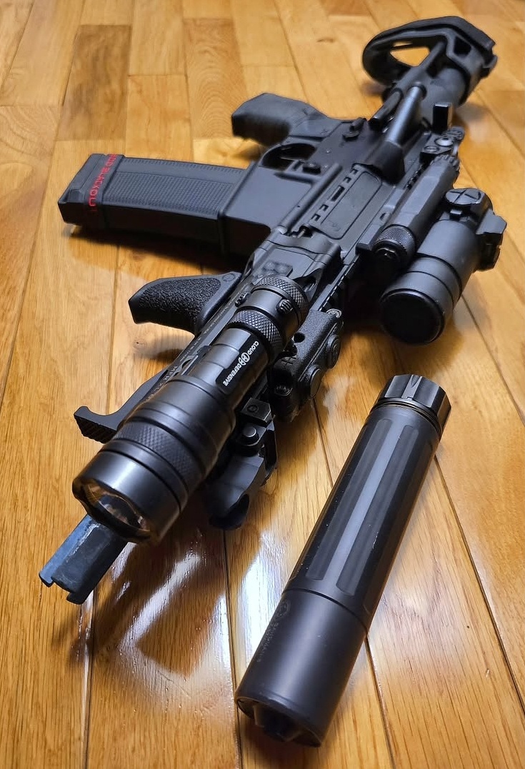 A short black AR-style pistol lying on a wood floor, with a weapon light on the handguard and a .300 Blackout magazine, and a Dead Air Sandman S suppressor lying on the floor in front of it
